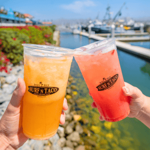 Two hands clink orange and red drinks in plastic cups labeled Surf N Taco at Baja Bay, set against a waterfront bustling with boats and a lively marina backdrop.