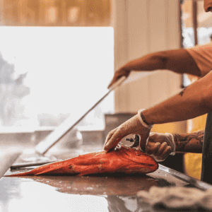 Wearing gloves, the person skillfully handles a large fish on a kitchen counter, knife in hand. Its evident that this cooking preparation scene is brought to life with the fresh offerings from Wild Local Seafood.