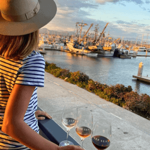 A person in a striped shirt and hat gazes over the marina from the Deep Sea Wine Tasting Room, holding a glass of wine. Three wine glasses sit elegantly on the ledge, capturing the essence of nautical indulgence.