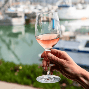 A hand delicately holds a wine glass filled with rosé, evoking the serene ambiance of a Deep Sea Wine Tasting Room, where boats and water merge into a dreamy blur in the background.