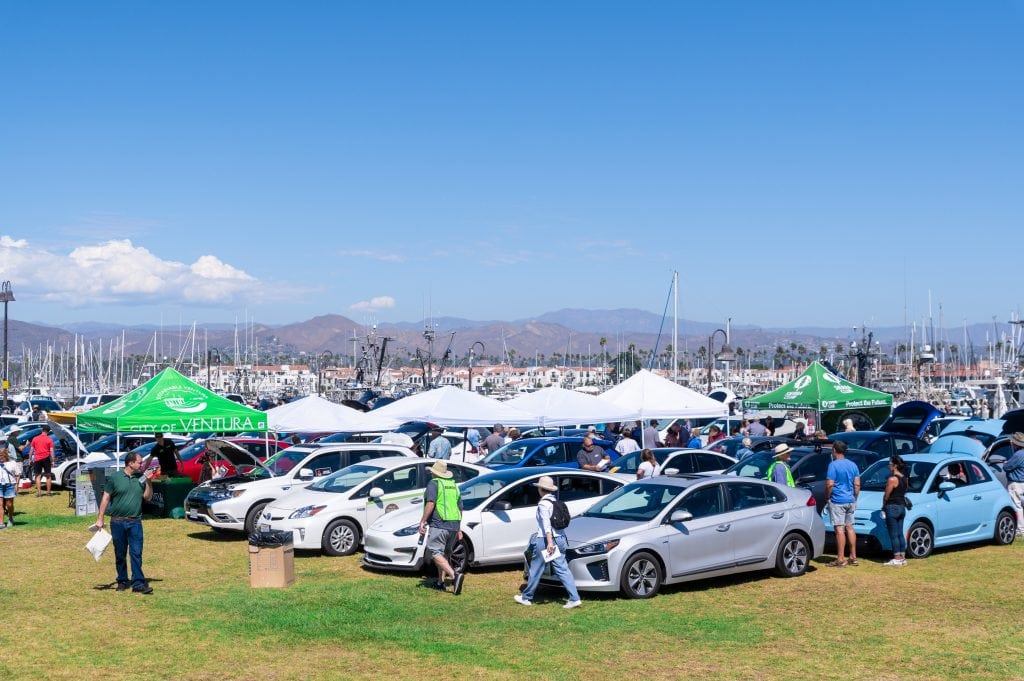 A busy outdoor event showcasing various electric and hybrid cars with attendees browsing the vehicles under multiple white tents on a clear day near a marina.