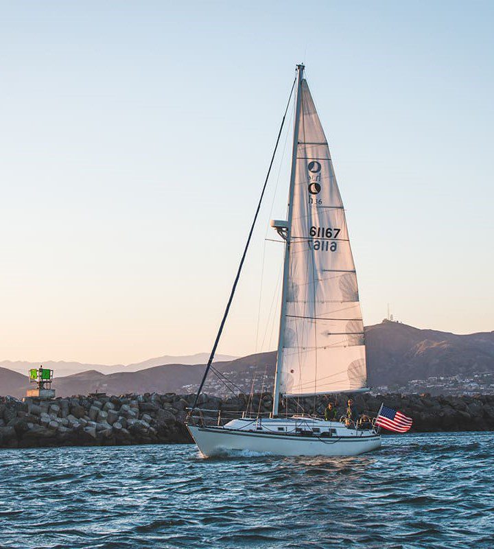 A sailboat with an American flag sails near a rocky breakwater during sunset; a green navigation marker and mountains are visible in the background.