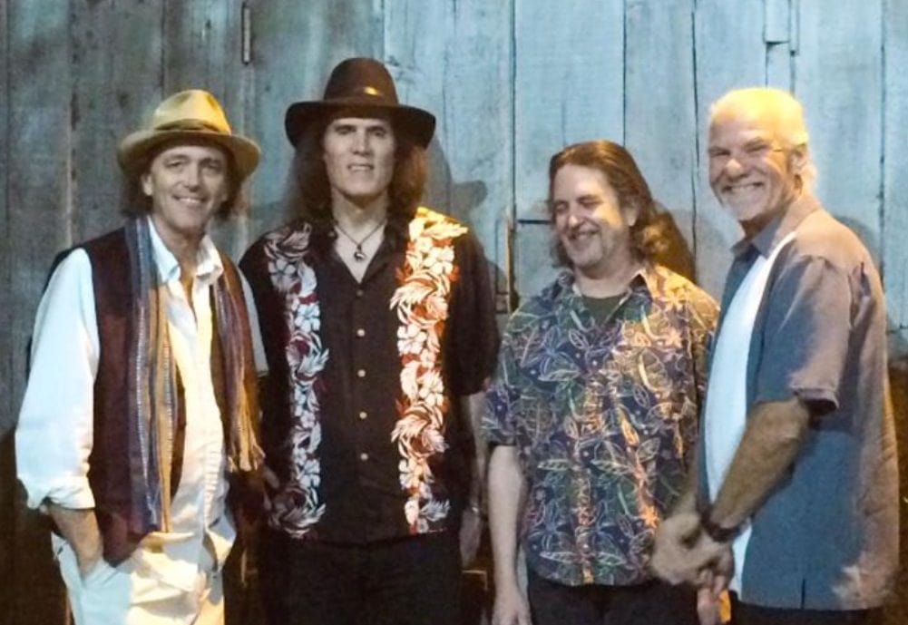 Four men standing together, posing for a photo in front of a wooden wall. They are dressed in casual attire with hats and patterned shirts.