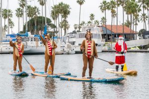 Three people in reindeer costumes and one in a Santa outfit paddleboard on a marina, with boats and palm trees in the background.