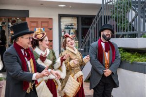 Four people in Victorian-style clothing sing joyfully outdoors, standing near a staircase.