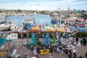 Harbor scene with festive boats decorated for the holidays, artificial snow falling, and people gathered along the waterfront.