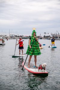 People in festive attire, including a person dressed as a Christmas tree and a dog in a holiday outfit, are paddleboarding on a calm body of water.