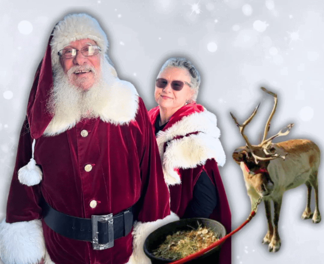Older man in a Santa costume and a woman in a red cloak stand beside a reindeer with snow in the background.