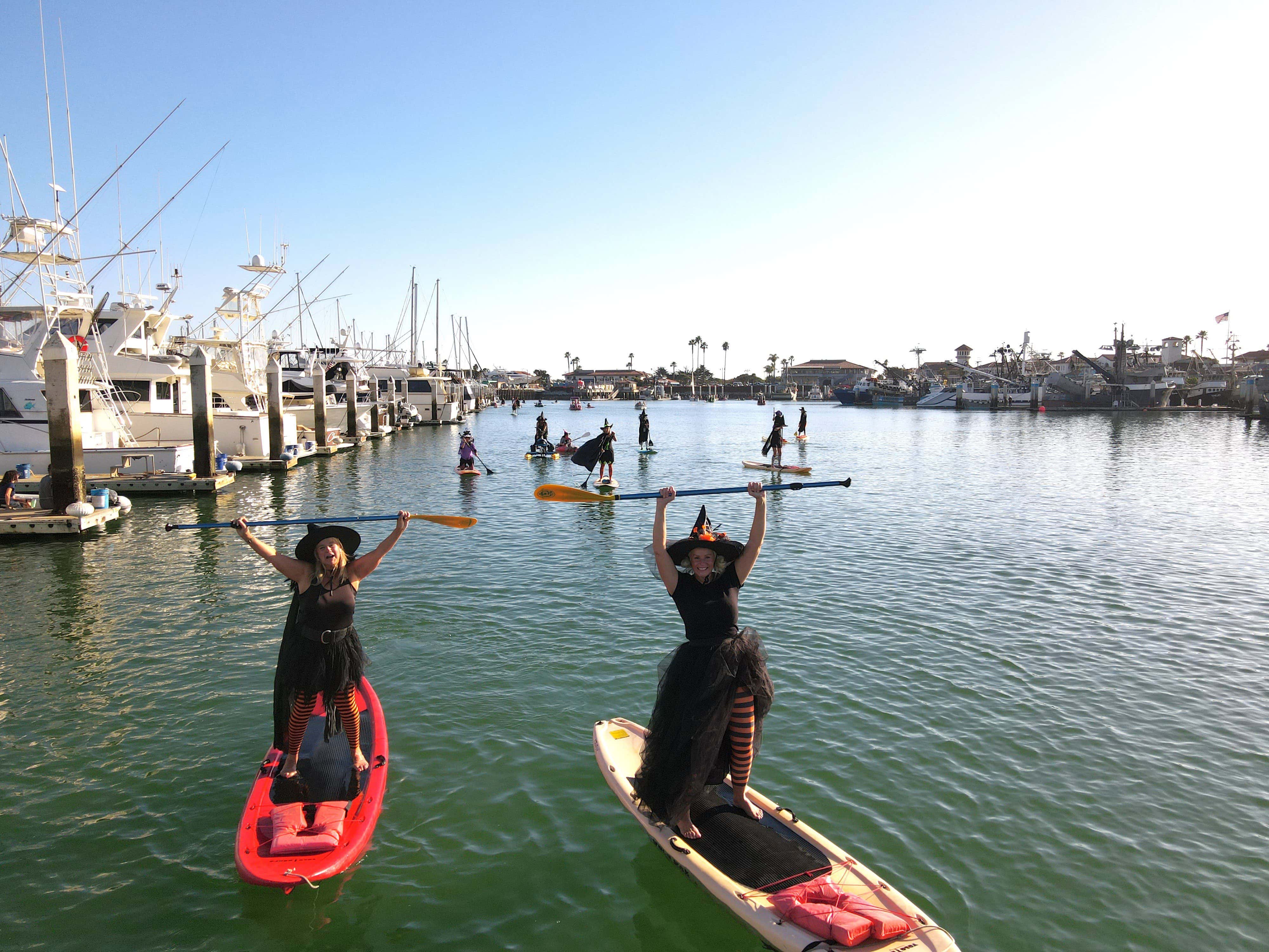 People dressed as witches paddleboarding on a calm marina, holding paddles overhead.