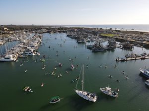 Aerial view of a marina with numerous boats docked and people paddling on the water. Surrounding area includes parking lots and coastline.