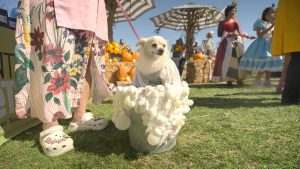 A small dog in a fluffy white costume sits in a bucket. Nearby, a person in floral attire and white clogs stands. Background includes pumpkins, people, and umbrellas on a grassy area.