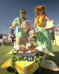 Two people in tropical outfits hold a dog with a blue ribbon. Another dog sits in a small decorated vehicle with the number 64. They pose outdoors on a sunny day.