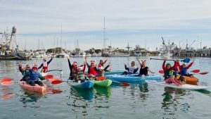 A group of people in colorful kayaks wave while floating in a marina. Boats and masts are visible in the background.