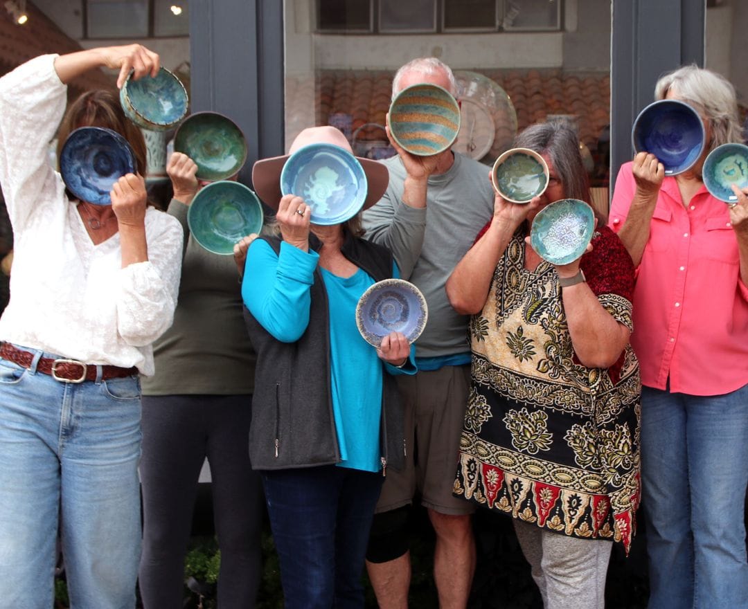 A group of people standing in front of a building, holding ceramic bowls up to their faces, each bowl displaying unique colors and patterns.