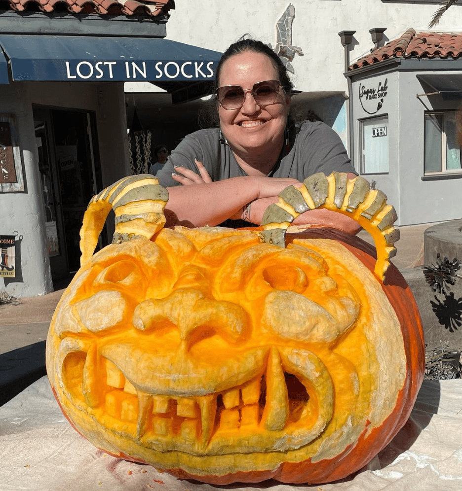 A person wearing sunglasses stands behind a large, intricately carved pumpkin with a smiling face.