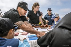 A group of people, including a child, gathered around a table on a boat, engaged in observing maps and taking notes.