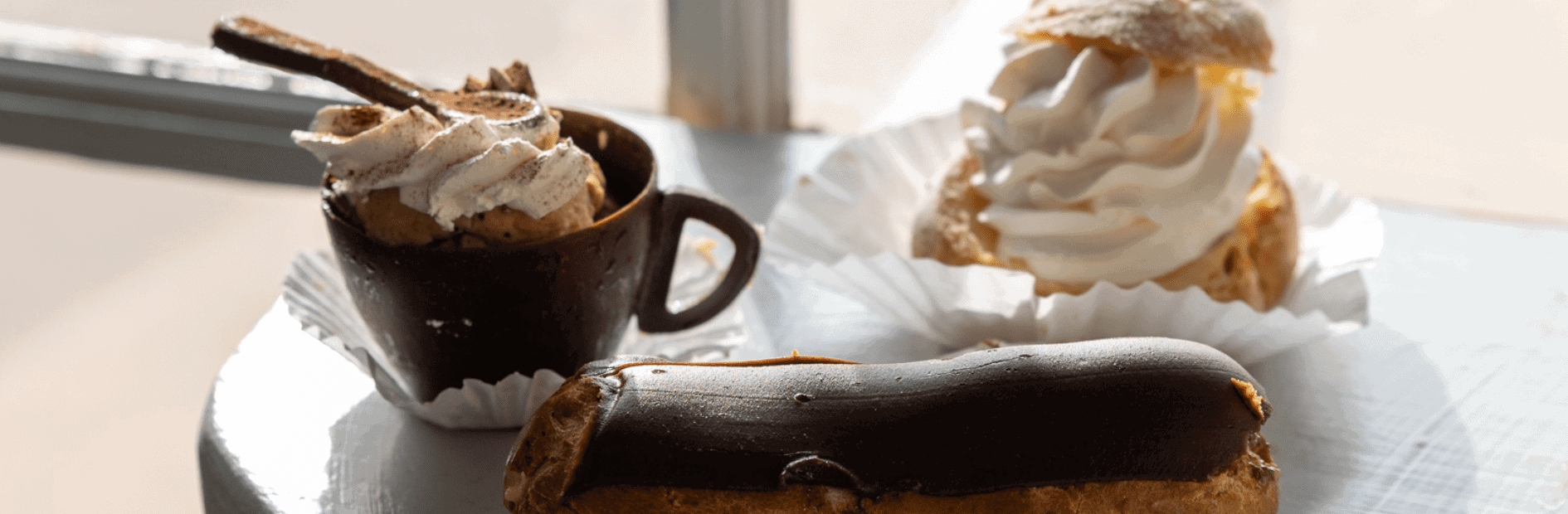 A cup of dessert with whipped cream, an éclair with chocolate glaze, and a cream puff on a table in natural light.