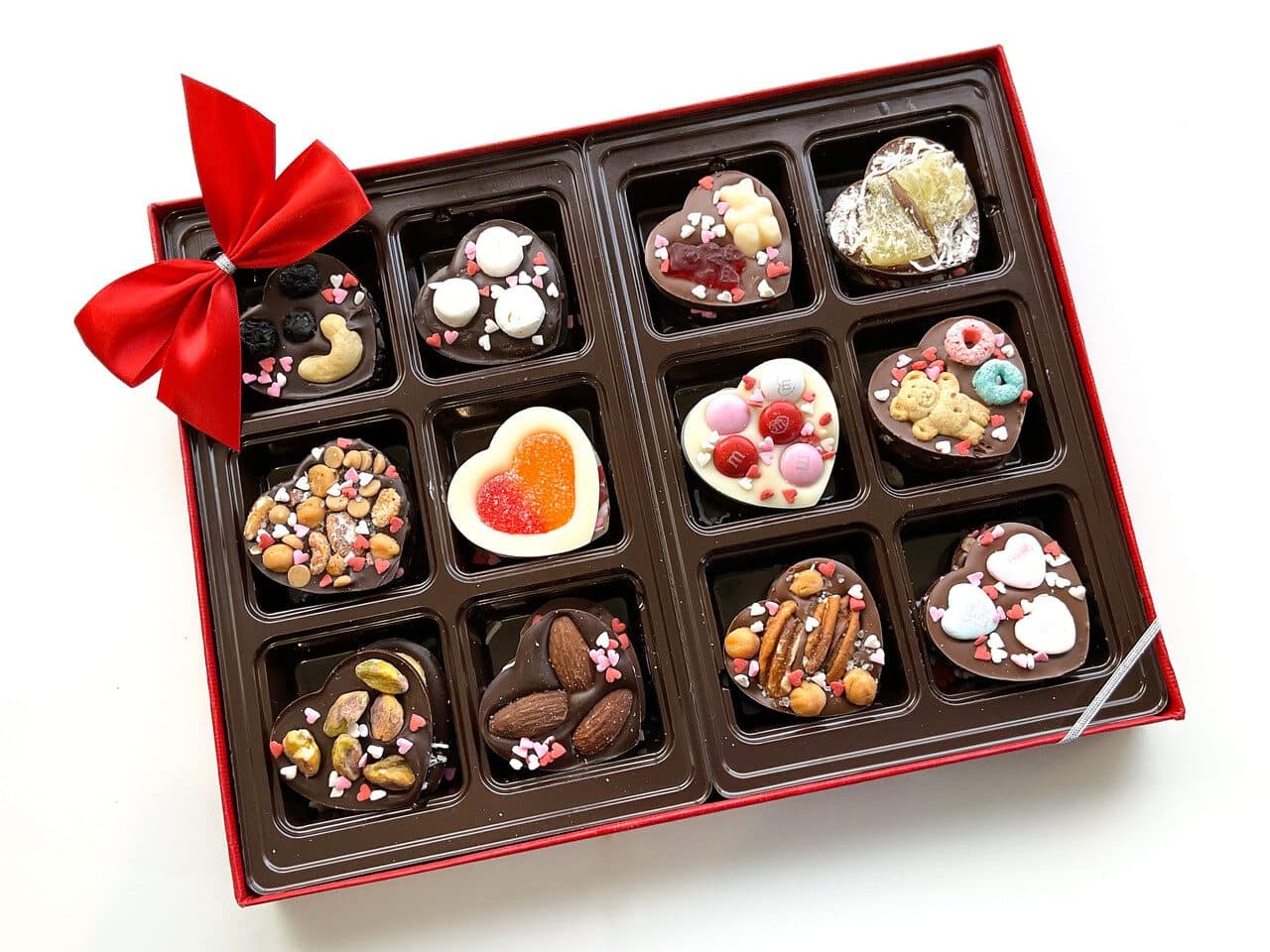 A box of 12 assorted decorated chocolates, each in its own compartment. Some chocolates have nuts, fruit, or candy toppings. The box has a red ribbon on one corner.