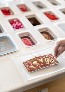 A hand with a glove decorates a chocolate bar with the words I ? YOU on it. Various toppings are visible in containers on the table.
