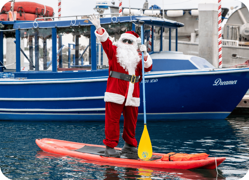 Santa Claus paddleboarding at Ventura Harbor Village during Christmas season.